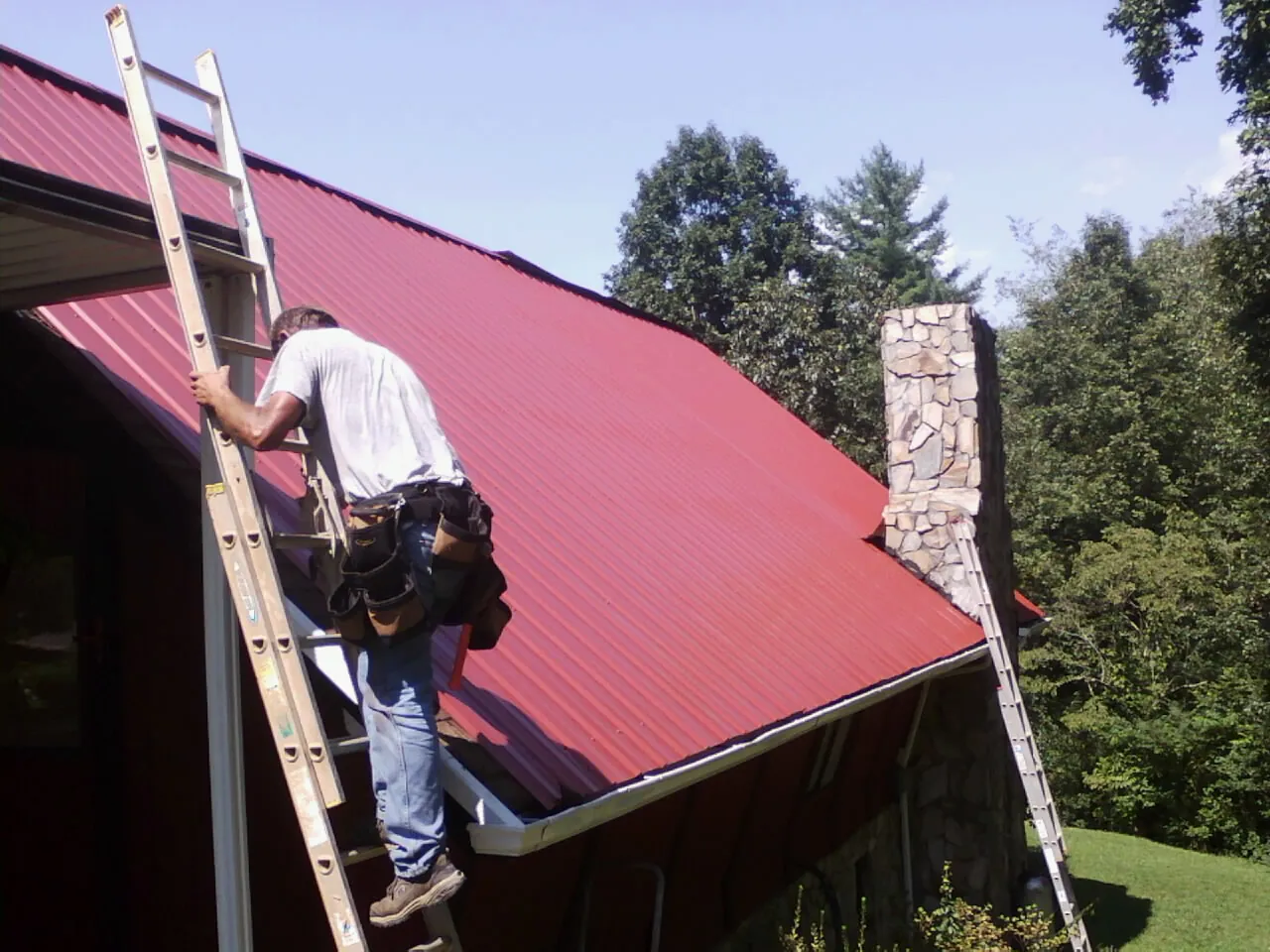 Roofer on a ladder beside a red metal roof and stone chimney — Soffit & Fascia Repair by Roofing Brothers in Wellesley